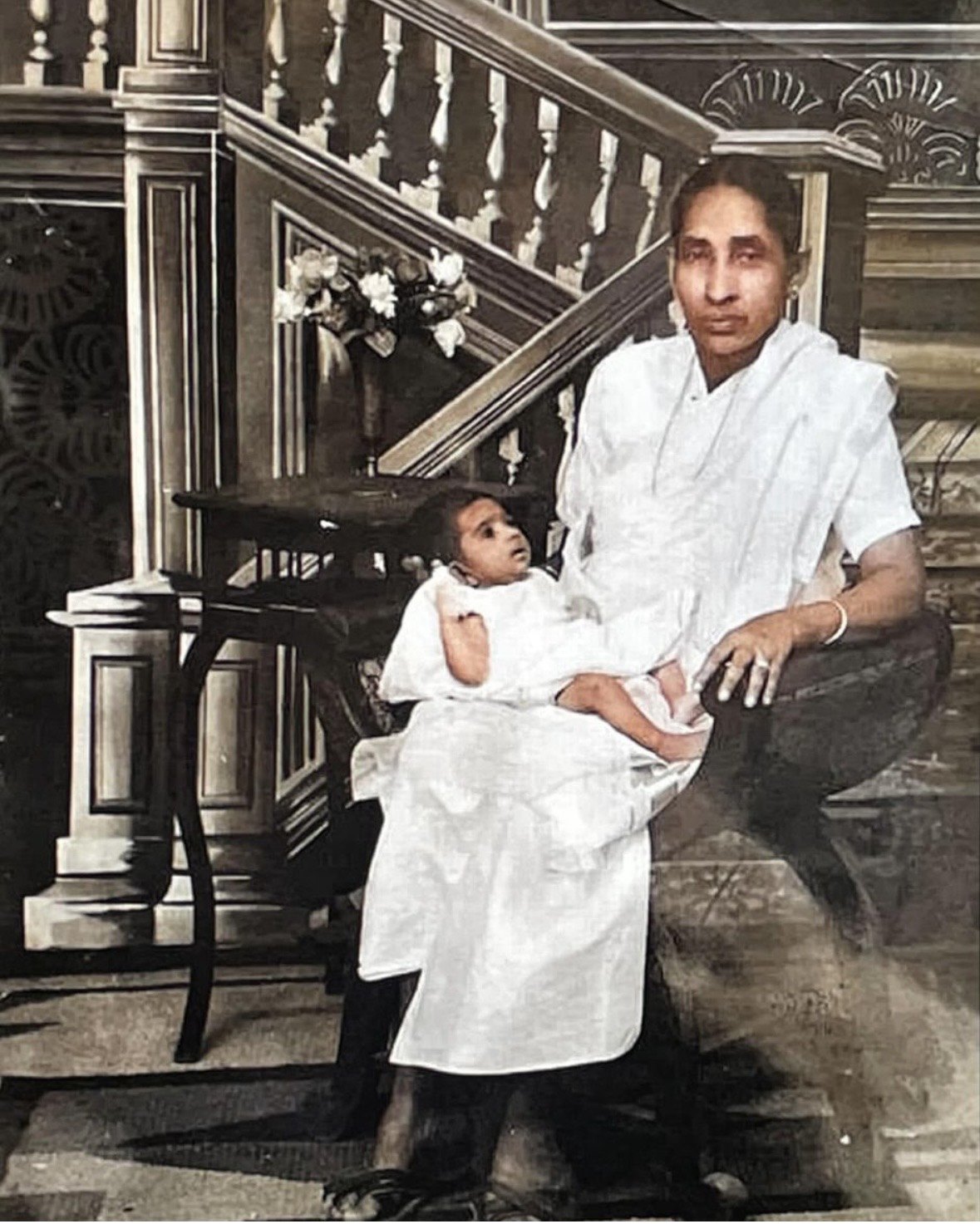 Grace's grandmother in white Kerala cotton, seated on a stool holding Grace as a month-old baby.