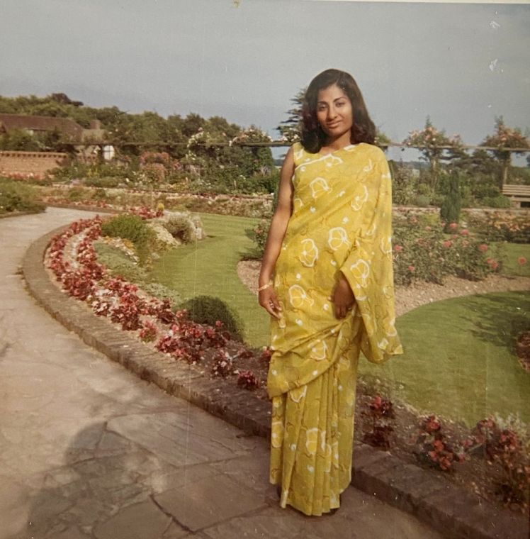 Grace in a yellow sari standing in a formal rose garden.